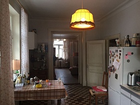  различные предметы на клетчатой коричневой клеенке обеденного стола у окна с настольной лампой на подоконнике кухни с бежевыми панелями на белых стенах