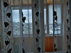 деревянный потолок и стены на застекленном балконе через белую гардину с рисунком на окне кухни яркой современной трехкомнатной квартиры с комнатой бабушки