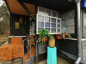 искусственные цветы под окном веранды и стол у лестницы с перилами на входе в деревянную избу в деревне