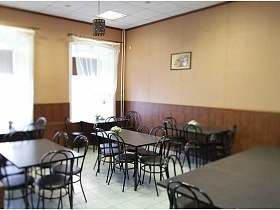 картина на бежевой стене с коричневыми панелями просторного зала с рядом темных столов со стульями типичной столовой
