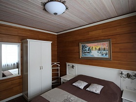 светильники на белой спинке большой кровати у деревянной стены с картиной в спальне съемного коттеджа