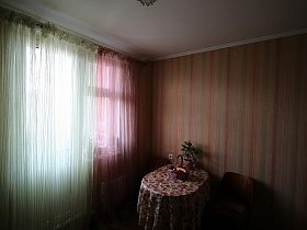 кухня с белой и розовой гардинами, полосатыми цветными обоями на стенах, цветной скатертью на обеденном столе типовой трехкомнатной квартиры неработающих пенсионеров