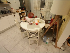плетенное кресло, белые стулья со спинкой вокруг белого обеденного стола в белой кухне с белой гардиной на окне современной трехкомнгатной квартиры