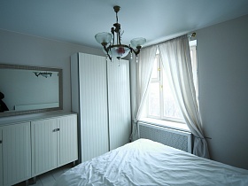 белый шкаф для одежды, белая высокая тумба у стены с прямоугольным зеркалом в рамке в светлой спальной комнате с белыми шторами на окне стильной современной квартиры