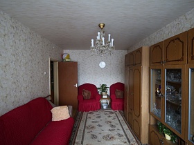 вишневые чехлы на мягкой мебели, мебельная стенка у светло-серых стен гостиной с цветным светлым ковром на полу и люстрой, стилизованной под свечи на белом потолке квартиры пенсионеров