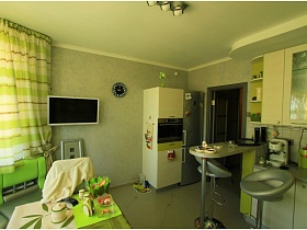 серебристый холодильник,белый жарочный шкаф и белый плоский телевизор на стене в зеленой кухне двухкомнатной квартире новостроя