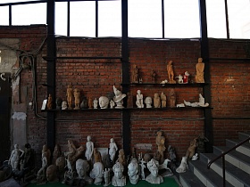 разнообразные скульптуры на полу, на полках у кирпичной стены рядом с серой лестницей складской комнаты тату салона в лофт стиле