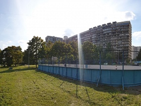 спортивная площадка за голубым забором с металлической сеткой поверх деревянных щитов на просторном зеленом участке между деревьями в жилом квартале Чертаново