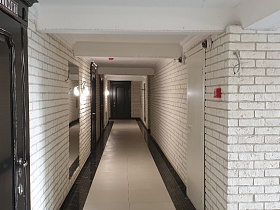 двухцветная плитка на полу длинного коридора с кирпичными стенами, темными входными дверьми в квартиры жилого дома