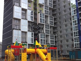 игровой комплекс с яркими красками на детской площадке внутри современных серых многоэтажек с цветными вставками и застекленными балконами новостройки