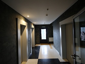 вид на современный ухоженный холл подъезда с темными стенами, белым потолком, квадратной плиткой на полу и двумя лифтами из открытой входной двери многоэтажного дома