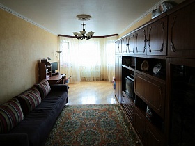 цветной ковер между темным мягким диваном и мебельной стенкой с телевизором в светлой гостиной песочного цвета