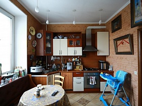 разнообразные кухонные принадлежности на столешнице бело коричневой мебели, диван у окна, круглый обеденный стол со стульями, детское кресло для кормления в кухне с одиночными подвесными светильниками на белом потолке в трехкомнатной советской квартире