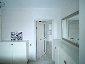 коллаж из фотографий на поверхности белого невысокого шкафа, белая тумба у стены с большим зеркалом в спальной комнате с открытой белой дверью