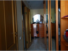 круглое зеркало в рамке над высоким деревянным коричневым комодом в прихожей трехкомнатной квартиры панельки