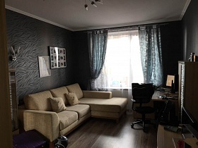 черное кресло у компьютерного стола с монитором, мягкий бежевый угловой диван с подушками у стен с темно серыми обоями, окна с серыми шторами и белой гардиной в спальной комнате современной квартиры