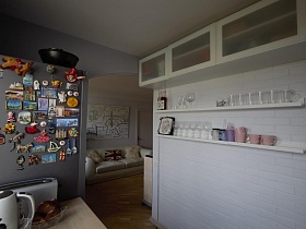 множество разнообразных магнитиков на поверхности серебристого холодильника, белый навесной шкаф с рифленным стеклом на дверцах, настенные полки со стеклянной посудой в светлой кухне стильной трешки