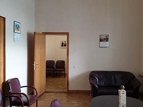 статуэтка на поверхности коричневого стола, вишневые кресла для посетителей, небольшой мягкий темнокоричневый диван у светлой стены с календарем у входной двери в приемную КГБ СССР для аренды для съемок кино