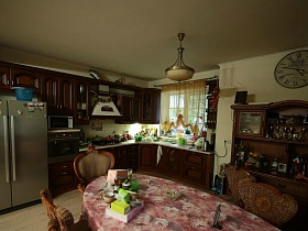 серебристый двухдверный холодильник у коричневой кухни, круглые часы над сервантом с посудой в просторной зонированной комнате семейного дома среди густого хвойного леса