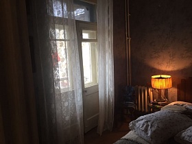  желтый абажур настольной лампы на стуле у большой кровати спальной комнаты с белой гардиной на балконной двери кв. 24