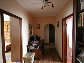 мягкий пуфик, письменный стол с монитором, принтером и компьютерным синим стулом в углу коридора с открытыми дверьми в разные комнаты трехкомнатной яркой квартиры жилого дома