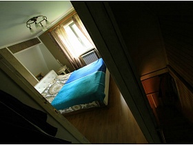 большая кровать с голубым покрывалом в спальной комнате со шторами на окне и плафонами круглой люстры через открытую дверь