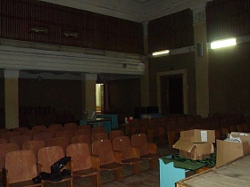 голубой стол с пустыми картонными коробками у деревянной сцены актового зала с колоннами, секционными креслами, деревянными решетками на желтых стенах старого клуба времен СССР