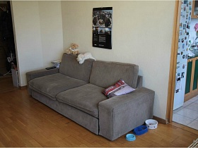 календарь над мягким серым диваном с подушками и мягким и игрушками на спинкев гостиной простой квартиры на Садовом