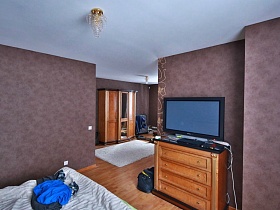 телевизор на комоде у коричневой стены напротив деревянной кровати спальной комнаты современного кирпичного дома