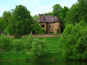 элитный кирпичный двухэтажный дом с крышей под черепицу, арочными окнами и балконом между колоннами за кирпичным забором на красивом берегу реки среди зелени в летнее время