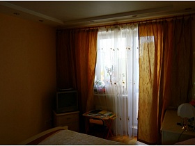 телевизор в углу на тумбе и детский столик у окна с ораньжевыми шторами в спальне приличной трешки панельного дома