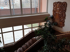 большие комнатные цветы в углу лестницы с деревянными перилами и барельефом на белой стене у окна столовой СССР