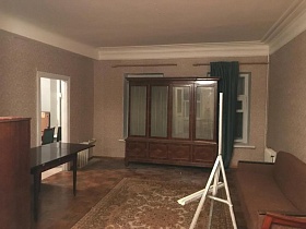 высокий коричневый четырехдверный шкаф со стеклянными вставками у белых окон с синими шторами, белый мольберт у коричневого дивана на полу гостиной с бежевыми стенами