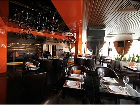 сервированные столики с индивидуальными креслами в ораньжевом зале евро ресторана с большими металлическими люстрами на натяжном потолке