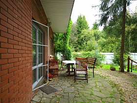 деревянный стол и стулья со спинкой на открытой площадке , выложенной диким камнем у входных дверей в семейный простой дом в глухом лесу