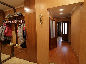 зеркальные дверцы шкафа-купе, коричневая мебельная стенка с одеждой на полках и крючках в прихожей трехкомнатной квартиры государственного служащего
