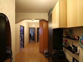 открытые полочки под бежевыми навесными шкафчиками, сумки на коричневом комоде, овальное зеркало на стене прихожей большой трехкомнатной квартиры в переезде