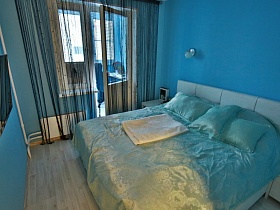 большая белая кровать с атласным покрывалом и подушками в спальне с голубыми стенами в молодежной евро квартире Новостроя