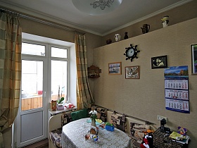 стол и угловой диван у бежевой стены с картинами, часами и перекидным календарем в светлой просторной кухне с желтыми полосатыми шторами на окне с балконной дверью семейной классической квартиры в сталинском доме