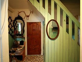зеркало в декоративной черной рамке с полочкой над креслом у входной двери под  деревянной лестницей с перилами и зеркалом в старорусской избе в деревне в деревне