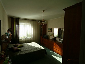 большая кровать с подушками и покрывалом, коричневые с зеленым шторы на окне светлой спальни квартиры оператора