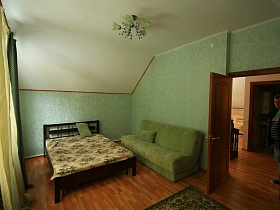 зеленый мягкий диван с подушками, деревянная кровать с цветным зеленым покрывалом в спальне с зелеными стенами и зеленой люстрой на потолке съемного двухэтажного дома в сосновом лесу