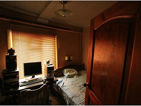 большая незаправленная кровать с подушкой, длинный стол с компьтерем и музыкальным центром у окна с закрытыми жалюзив спальной комнате дачи через открытую дверь
