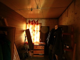 красная штора на окне комнаты с вещами, сломанной мебелью и разным хоз инвентарем в небольшом домике отшельника среди новостроек