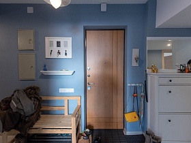 прямоугольное зеркало над белым высоким комодом, деревянная скамейка с вещами, обувь на коврике, зонтик и сумочки на деревянной вешалке у входной двери в голубую прихожую современной сталинской квартиры