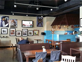 плоский телевизор и разнообразные картины на светлой стене , напольная деревянная вешалка и посудомоечное отделение с голубой плиткой в зале кафе в темных тонах