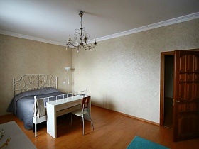 стул со спинкой, белый деревянный стол у белой кровати с металлической резной спинкой и синим покрывалом в светлой спальной комнате квартиры в переезде(въезде) молодоженов