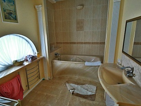 зашитая бежевая ванна у стены с бежевой плиткой, зеркало в рамке над бежевой раковиной, полотенцесушитель под арочным окном в ванной комнате художественной дачи
