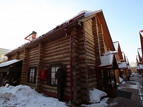 внешний вид деревянных бревенчатых домиков из сруба с деревянными ставнями на окнах, крышей над открытым крыльцом и навесом над мангалом