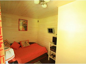 ноутбук на белой этажерке, телевизор на стене над разложенным диваном с красным покрывалом и подушками, белый абажур настольной лампы на тумбочке в спальной комнате современной дачи работника кино